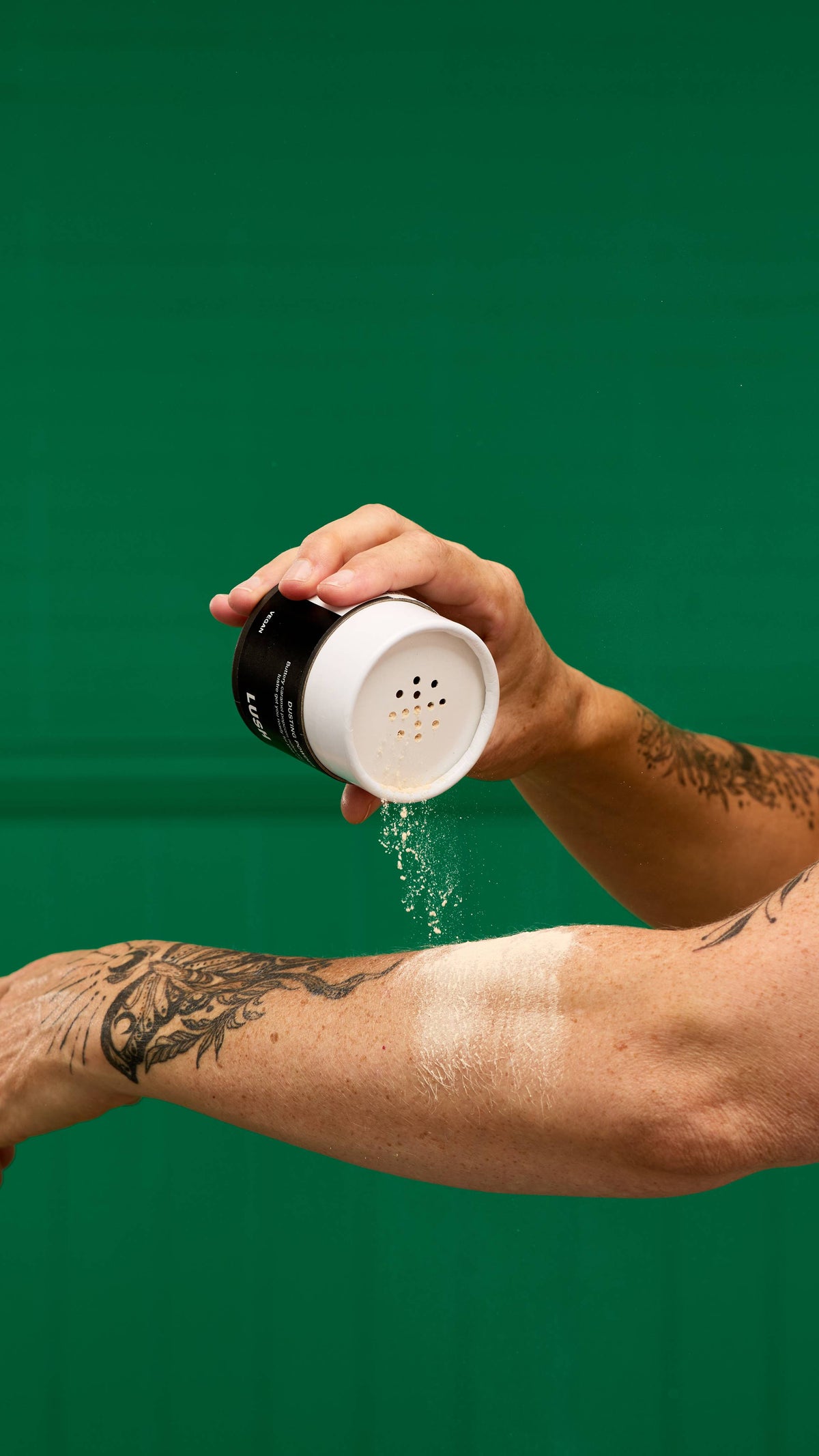 A person's hand shakes a LUSH Let the Good Times Roll dusting powder container, with a fine cloud of white powder falling onto their tattooed forearm. The background is a vibrant green.