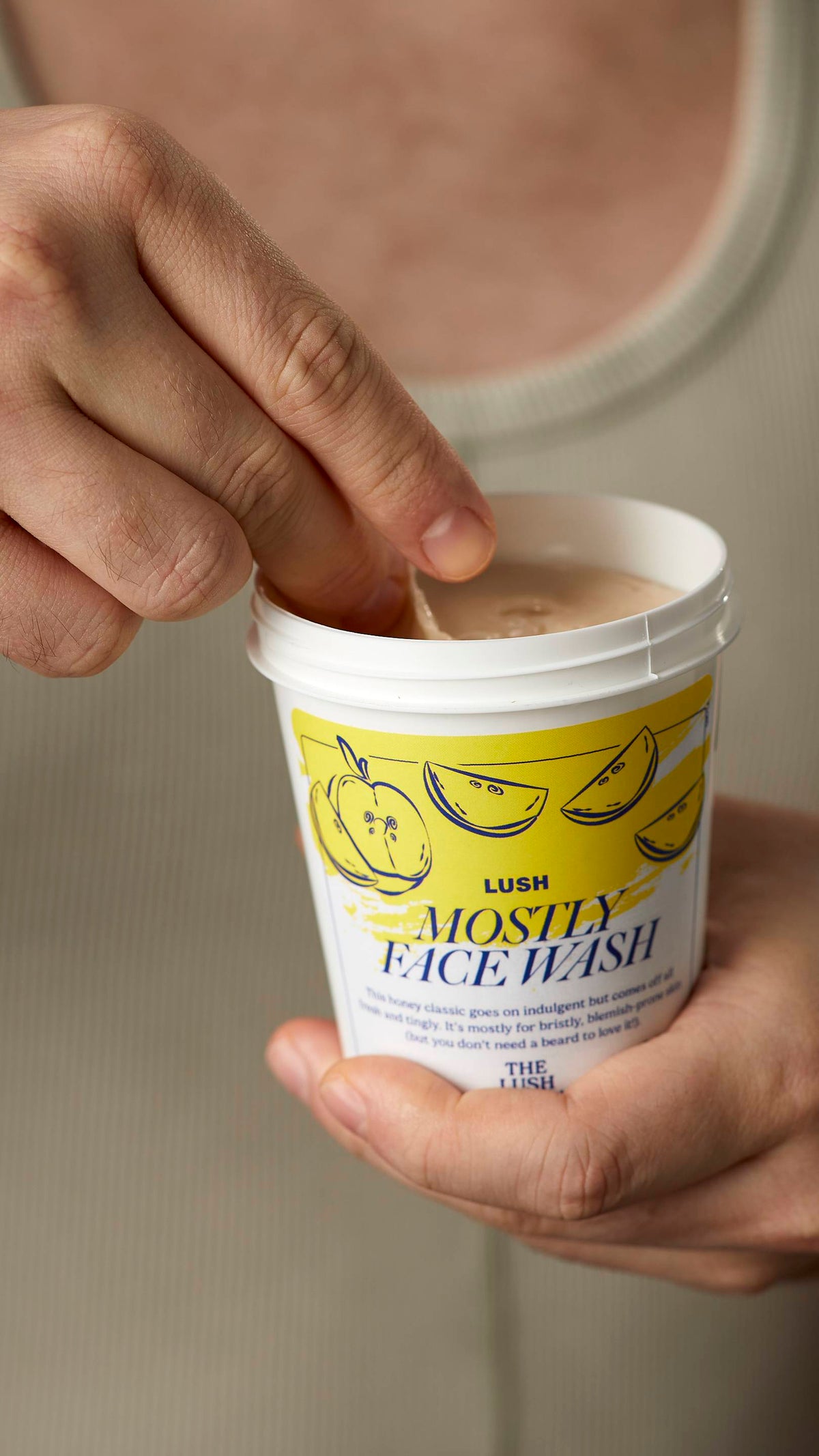 A close-up image of the model's hands. One hand holding the white, LUSH version-themed packed pot of the Mostly facial wash and scooping out the product with the other hand.