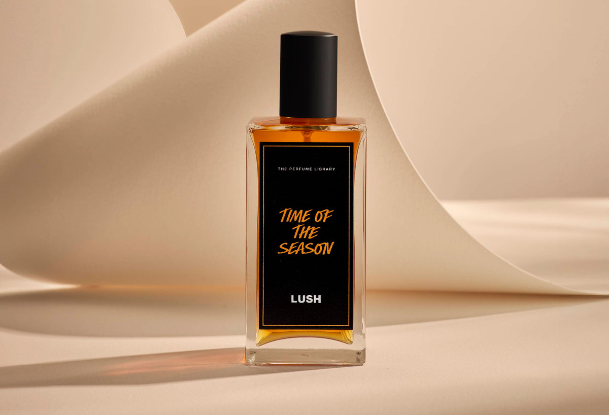 The image shows the Time Of The Season glass perfume bottle. It is sitting on a soft, beige background with curved, cream paper creating soft, gliding shapes behind the product.